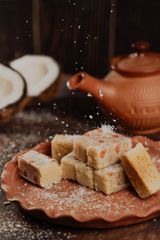 Traditional Indian coconut burfi sweets on a terracotta plate, with shredded coconut sprinkled around and a clay teapot and coconut halves in the background.