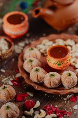 Festive Indian sweets shaped like small pumpkins, coated in coconut, arranged on a terracotta plate with decorative clay pots, cashews, rose petals, and a rustic clay teapot in the background.