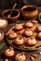 Round Indian sweets topped with whole almonds in red paper liners on a golden tray, surrounded by earthen bowls and honey in the background.