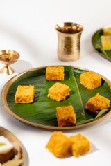 Bright orange squares of Indian sweet on a banana leaf–lined plate, with a brass lamp and cup in the background.