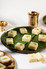 Squares of white Indian barfi topped with crushed pistachios on a banana leaf–lined plate, with a brass lamp and cup in the background.