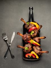 Skewers of chicken lollipops and colorful bell peppers on a metal tray with a red napkin, knife, and fork