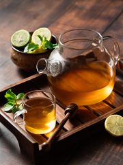 Herbal tea in a glass teapot and cup with lemon slices, mint leaves, and honey dipper on a wooden tray