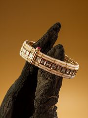 Luxurious gold bracelet with square-cut stones and delicate pearl details displayed on a dark wooden hand sculpture against a warm golden background.
