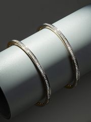 Pair of sleek gold bangles embellished with baguette diamonds, displayed on a smooth cylinder against a dark grey backdrop.