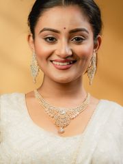 Smiling woman in a white traditional outfit wearing an elegant diamond and blue gemstone necklace with matching earrings, set against a warm golden background.