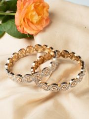 Gold bangles with intricate circular diamond settings placed on a light fabric near an orange rose.