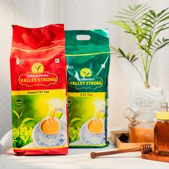 Two vibrant packs of Sinolacherra Valley Strong and Goombira’s Valley Strong Assam CTC Tea displayed with a honey jar and potted plant on a wooden surface, highlighting garden-fresh quality.