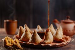 Plate of crispy, golden samosas arranged in a circle, with tamarind sauce being drizzled onto one samosa, and a clay teapot and steaming cup in the background, creating a warm and inviting scene.