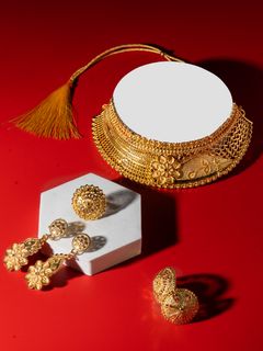 A gold choker necklace with a tassel, matching earrings, and a ring displayed on a red background alongside a white hexagonal stand.