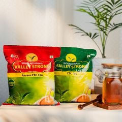 Two vibrant packs of Sinolacherra Valley Strong and Goombira’s Valley Strong Assam CTC Tea displayed with a honey jar and potted plant on a wooden surface, highlighting garden-fresh quality.