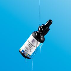 A sleek bottle of face serum with a dropper, displayed on a white pedestal against a vibrant blue background.