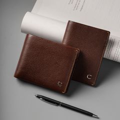 Two premium brown leather wallets placed on a gray surface, accompanied by a pen, showcasing a sophisticated handcrafted design.