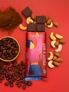 A partially unwrapped Fancis Fruit & Nut chocolate bar on a red background, surrounded by coffee beans, ground coffee, and cashews.