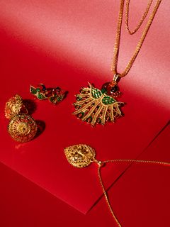 A gold necklace with a fan-shaped pendant featuring green enamel, displayed alongside gold filigree earrings and a teardrop pendant on a red background.