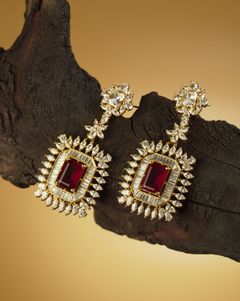 Opulent gold earrings featuring deep red center stones and sparkling diamonds, displayed on a dark wooden surface against a warm golden backdrop.