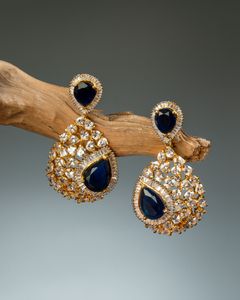 Exquisite teardrop gold earrings adorned with deep blue gemstones and sparkling diamonds, displayed on a wooden branch against a soft gradient backdrop.
