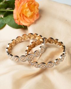 Gold bangles with intricate circular diamond settings placed on a light fabric near an orange rose.