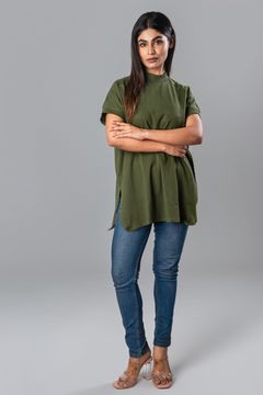 Woman crossing her arms while wearing a green high‐neck tunic top with short rolled sleeves, styled with fitted jeans and clear heels.