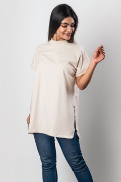A woman wearing a short-sleeve beige top with side slits and a high neckline, paired with jeans on a neutral background, showcasing a casual style.