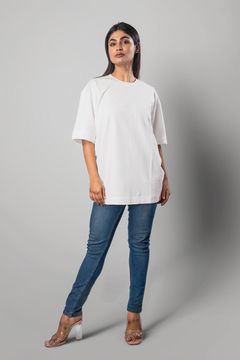A woman wearing a white oversized short-sleeve top with a relaxed fit, paired with jeans and heels on a neutral background, showcasing a casual modern style.