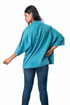 A woman wearing a loose teal top with wide sleeves, viewed from the back, paired with jeans on a white background, showcasing a relaxed casual style.