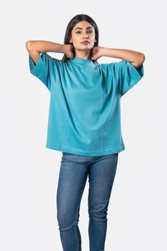 A woman wearing a loose teal top with wide sleeves, arms raised behind her neck, paired with jeans on a white background, showcasing a relaxed casual style.