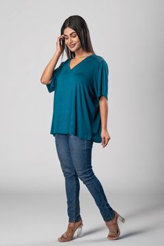 A woman wearing a teal loose-fit V-neck top with lace shoulder detail, paired with blue skinny jeans and clear-heeled sandals, posing against a neutral gray background.