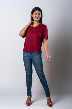 A woman wearing a maroon V-neck short-sleeve top, blue jeans, and open-toe brown heels, posing against a grey background.