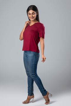 A woman wearing a maroon V-neck short-sleeve top, blue jeans, and transparent heels, posing against a grey background.