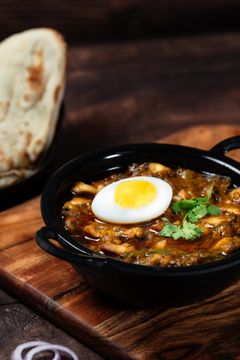 A bowl of stew with a hard-boiled egg and a slice of bread on a wooden cutting board