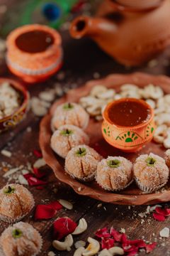 Festive Indian sweets shaped like small pumpkins, coated in coconut, arranged on a terracotta plate with decorative clay pots, cashews, rose petals, and a rustic clay teapot in the background.