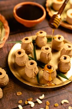 Earthen pot–shaped Indian sweets arranged on a golden tray, with honey being drizzled from a wooden dipper and scattered nuts in the background.