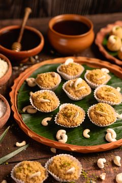 Small Indian sweets in paper liners, topped with cashews and pistachio crumbs on a banana leaf–lined clay plate, with earthen pots and scattered nuts in the background.