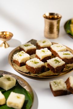 Two-layered Indian sweet squares topped with crushed pistachios on a golden tray, with a brass lamp and cup in the background.