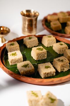 Squares of Indian barfi garnished with crushed pistachios on a banana leaf–lined clay plate, with a brass lamp and cup in the background.