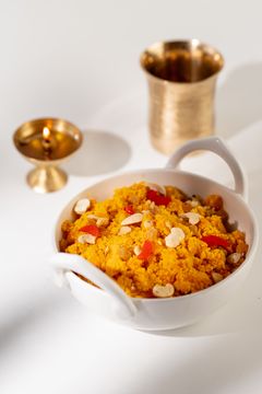 A white bowl with handles containing bright orange Indian sweet garnished with cashews and dried fruit, with a brass lamp and cup in the background.
