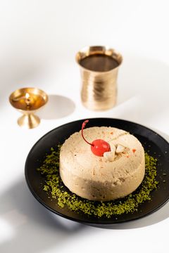 A round Indian sweet topped with a bright red cherry and cashews on a black plate sprinkled with crushed pistachios, with a brass lamp and cup in the background.