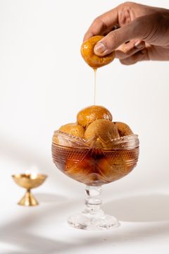 A hand holding a syrup-dripping gulab jamun over a bowl of golden gulab jamuns, with a small brass lamp in the background.