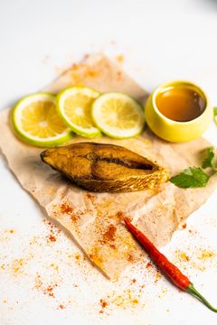 Close-up of a fried fish slice on parchment paper, garnished with fresh lemon slices, chili pepper, and a small bowl of sauce, showcasing a spicy Indian seafood dish.