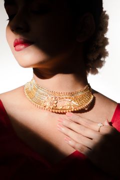 A woman in a red blouse wearing an ornate gold choker necklace and a ring on her hand, with her hair adorned with flowers, captured in soft, dramatic lighting.