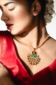 A woman in a red blouse wears a gold necklace with a green fan-shaped pendant, her hair adorned with flowers, shown against a bright background.